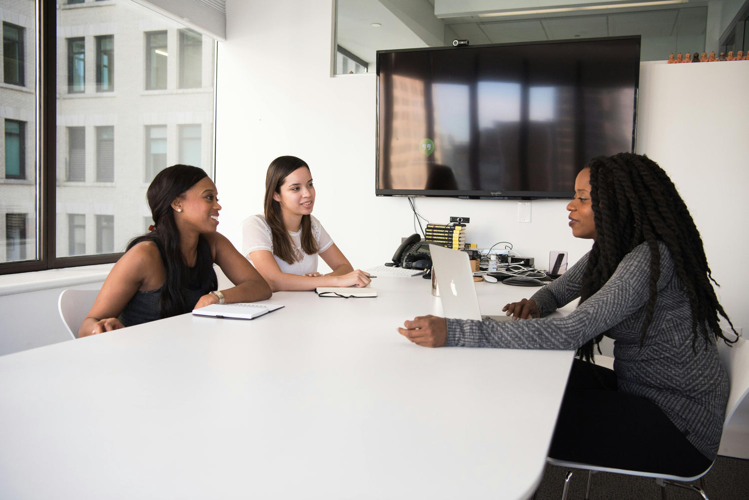 Three women in a meeting room with a TV screen in the background.